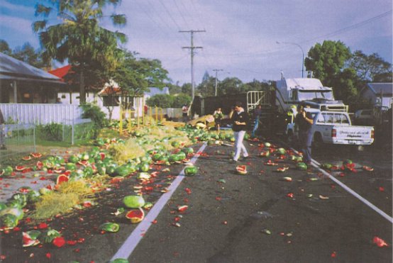 There is an accident in which watermelons are strewn over the road.  the road is blocked by a truck and an ute
