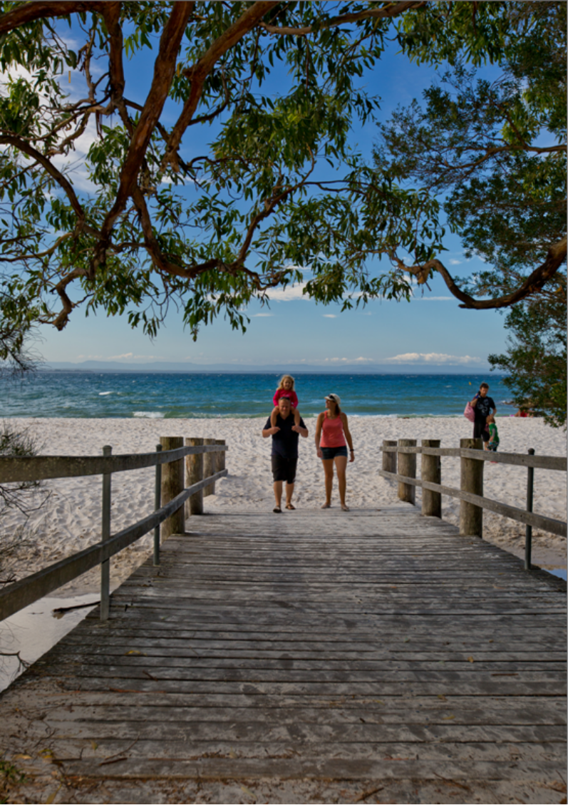 Photograph of a family leaving Murrays Beach in Booderee National Park, about to cross a wooden footbridge - with the ocean appearing in the background.