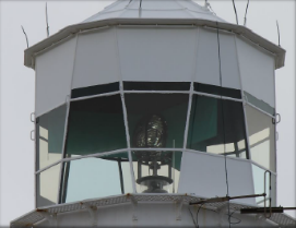 Photograph showing white lighthouse lantern room with glazing panes arranged around lantern room. A small lens can be seen through glazing inside the lantern room.