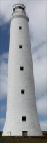 Photograph showing tall, white, stone lighthouse tower with lantern room on top. Five windows are lined vertically down the tower, and a door can be seen fixed to the base of the tower.