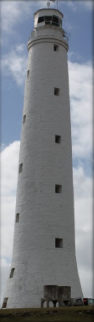 Photograph showing tall, white, stone lighthouse tower with lantern room on top. Ten windows are lined down the tower, and a door can be seen fixed to the base of the tower. Two water tanks can be seen close to the base of the tower.
