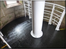 Photograph showing black, round iron floor. Curved stone wall encases floor, with white tube fitted to centre of floor. White railing pictured behind tube.