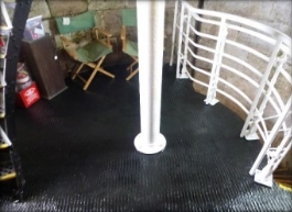 Photograph showing black, round iron floor. Curved stone wall encases floor, with white tube fitted to centre of floor. White railing and green chairs pictured behind tube.