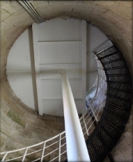 Photograph showing internal face of round, stone room. White underside of round floor above supported by while pole and floor beams. Black spiral stairs leading up to the white floor.