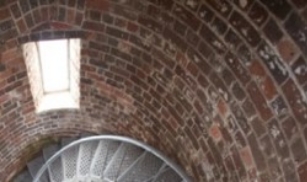 Photograph of curved, internal brick wall of lighthouse. Rectangular window is set into brick wall. Curved lattice staircase wraps around wall to lower floor. 
