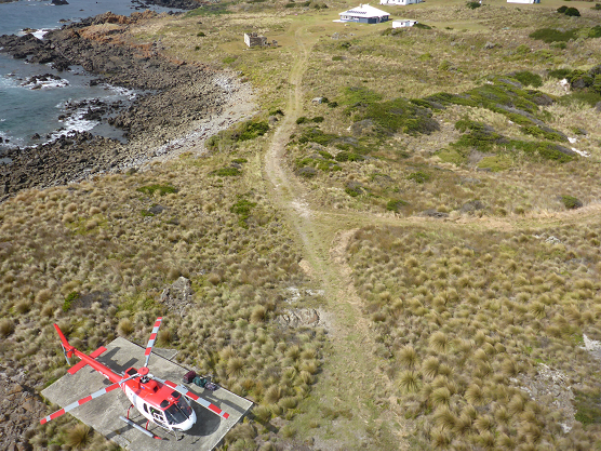 Photograph showing aerial view of coastal landscape. A red and white helicopter rests on a concrete square on the ground.