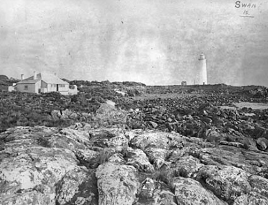 Black and white photograph showing rocky landscape. A white lighthouse tower and keepers cottage stand in distance. 