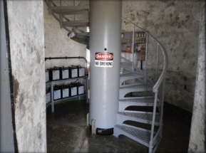 Photograph showing ground floor of lighthouse. One large central column is fixed to centre of room with a spiral staircase wrapped around it. 