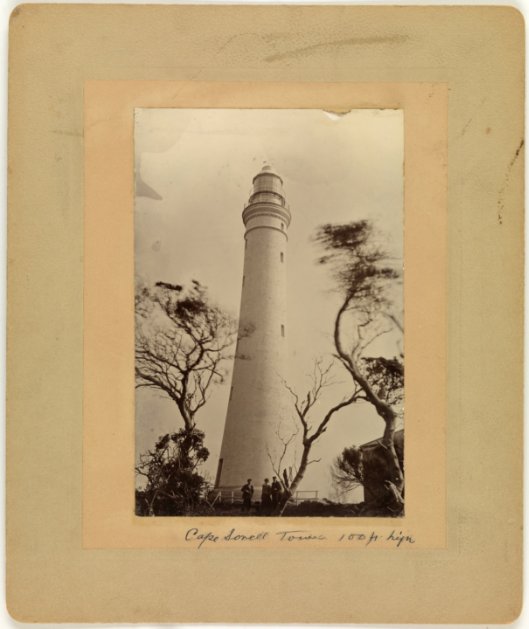 An old photograph of a white lighthouse tower with three figures standing at its base. Pencil writing below reads "Cape Sorell Tower 100ft high"