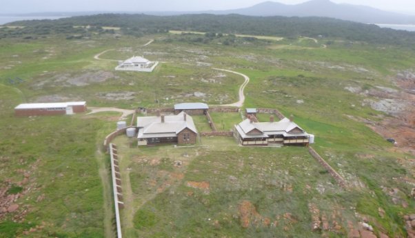 Photograph of collection of buildings within a rocky, vegetated landscape. Buildings include stone keepers cottages, brick service buildings, and several windbreak walls.