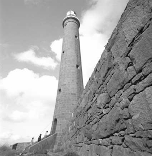 Black and white photograph of tall granite lighthouse tower beside a windbreak wall. Three figures are walking up the external steps into the tower. 