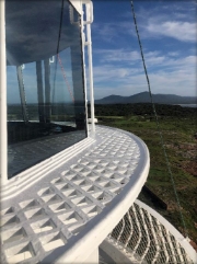 Photograph of white lantern house with white lattice catwalk curved around external edge of house, above a set of glazing panes. Mountains can be seen in distance.