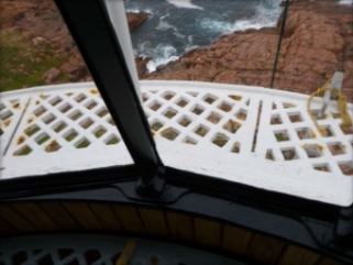 Photograph of white lattice external catwalk as seen through glazing panes inside lantern house. A rocky cliff edge can be seen below.