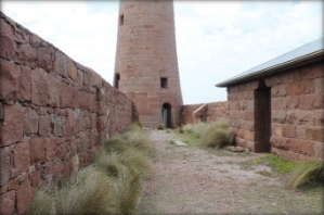 Photograph of base of granite lighthouse tower. A windbreak wall runs to side of tower, and a service building sits on its righthand side.