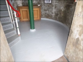 Photograph of round floor inside lighthouse tower. The base of a winding staircase and a green column are fitted to floor.
