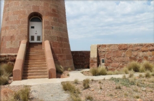 Photograph of granite lighthouse base with entry steps leading to double doors. A white path leadings from the bottom of the steps and past a granite windbreak wall.