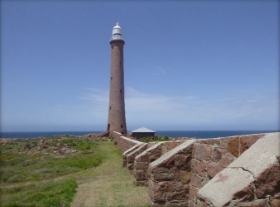 Photograph of tall granite lighthouse tower against blue sky. A long granite windbreak wall runs from the base of the tower out of sight.