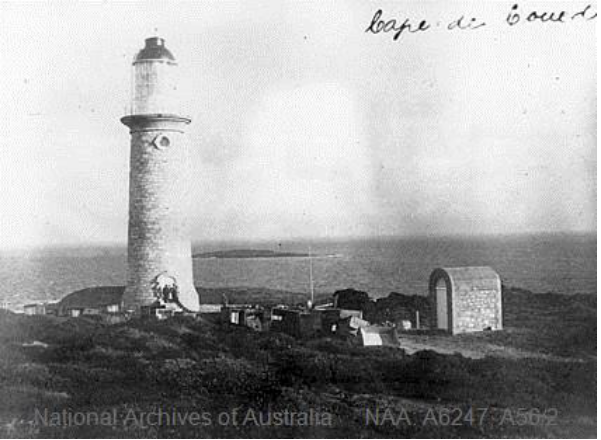 Black and white photograph of stone lighthouse tower surrounded by low-lying vegetation. A white, arched shed stands a little distance away from the tower, and the sea can be seen behind.