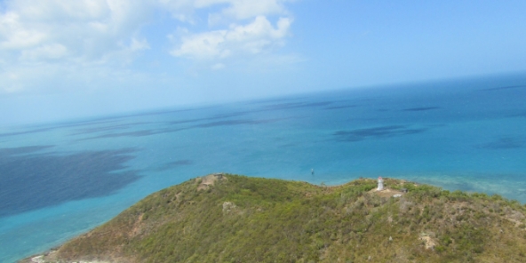 Photograph showing a small green island in a vast ocean. A small white lighthouse stands at the highest peak of the island.