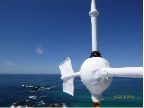 Photograph showing close up of wind vane. Blue ocean and rocky outcrops can be seen below in background. 