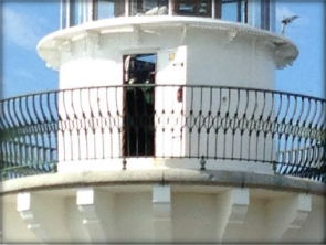 Photograph showing lighthouse balcony with gunmetal railing. The lantern house door stands ajar. 