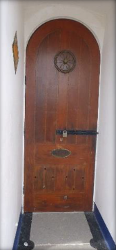 Photograph showing arched wooden entrance door with large sliding bolt lock and round vent fixture.