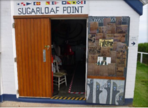 Photograph showing open wooden double doors of small white building. Painted above the doors is a collection of navigational flags and the words "Sugarloaf Point".
