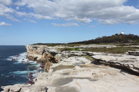 Photograph of coastal cliff edge beside a blue ocean. A small white lighthouse can be seen in the distance amongst green vegetation. 