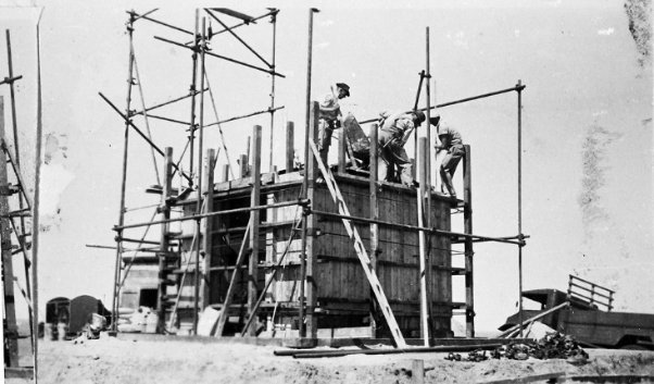 Black and white photograph showing persons building the ground floor of lighthouse tower surrounded by scaffolding and materials.
