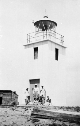 Black and white photograph showing a group of people standing at the base of a small white lighthouse tower.