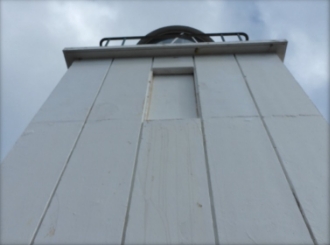 Photograph showing one side of a lighthouse tower with rectangular window cut into the concrete wall. The window is filled in with cement sheeting.
