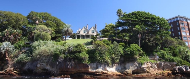A picture of Kirribilli House and garden from the harbour displays the large trees, bushes and rocks that surround the houses parameter. 