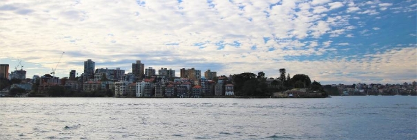 A picture overlooking North Sydney from the Opera House containing residential buildings, skyscrapers and Kirribilli House across a body of water and surrounded by blue skies.