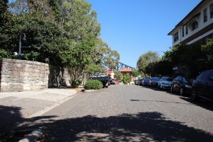 A picture of Kirribilli Avenue from street view showing a small portion of harbour bridge, a stone wall on the left side marking the property's boundary and parked cars across to the right side.