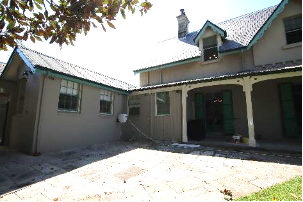 A continuation picture of Kirribilli House showing more of its south elevation, with smaller windows on the bottom floor, an entry doorway and stone pavement. 