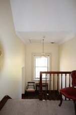 A picture of a stairwell landing at Kirribilli House with one chair to the right and an interim landing below with a chair, table, hanging light and medium sized window.  