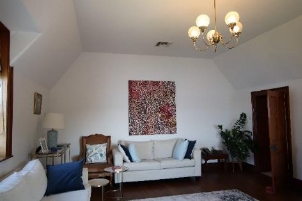 A picture of Kirribilli House family room with two white sofas, medium sized floor plant, table with a lamp and picture framed, art on the main wall and wooden floor, doorway and window frames.