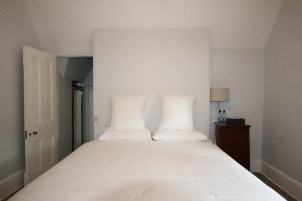 A picture of bedroom 4 at Kirribilli House with white walls, door, bedsheets and a wooden cabinet with a lamp to the right.