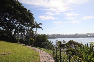 A picture of a grass area overlooking the harbour with a stone path and metal fence marking a boundary between the grass lands and below bushes and trees surrounding the harbour
