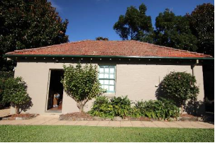 A picture of a garage at Kirribilli House with beige walls, green framed windows and orange brick roof with small trees and bushes along the side. 