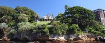 A picture looking at Kirribilli House from the harbour depicting the property perched up on the edge of the harbour and surrounded by large trees and bushes. 