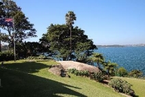 A picture of a Kirribilli House hilled garden with a large rock between the top and bottom hill, Australian flag pole to the left and surrounding grass lands, trees and water. 