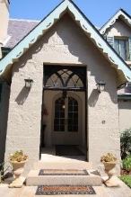 A close up picture of the Kirribilli House porch and entry way showing the triangular roof, stone walls, two ceramic vase plants and outdoor mats on the two steps leading to the arched entry door.