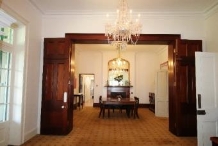 A picture of the Kirribilli House dining room with the dark wood stained doors open, showing matching dining table and chairs and, warm light chandeliers from the ceiling 