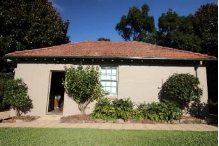 A picture of the Kirribilli House garage showing red brick roof, beige walls, green framed windows and with small trees and bushes outside. 
