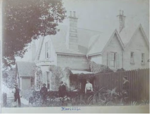 A black and white picture of 5 people standing in front of the Kirribilli House fence, beside large plants and the houses side, exposing the two chimneys and opened windows