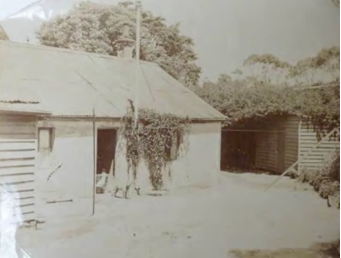 A sepia toned picture of small buildings with slated roofs, panelled walls and surrounded by trees 