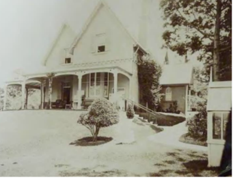 A black and white image of Kirribilli House from slightly below the sloped lawn showing the house's structure, a small singular tree and outdoor furniture