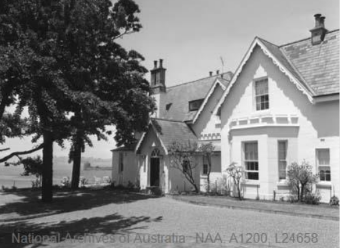 A black and white image of Kirribilli House entryway and driveway with two large trees by the entry of the house, opened windows and shaded lawns