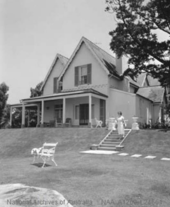 A black and white image of Kirribilli House from slightly below the sloped garden, showing the house's structure, a person walking down the garden stairs and outdoor furniture